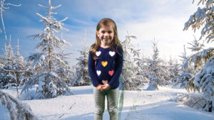 girl wearing hearts sweater standing in snow with snow covered trees background