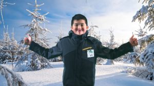 boy in a winter coat with his tongue out and two thumbs up with winter trees