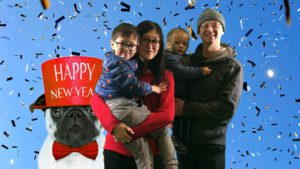 large pug dog wearing a red bow tie and red top hat that says happy new year, woman hold a child, man holding a baby with confetti in the background
